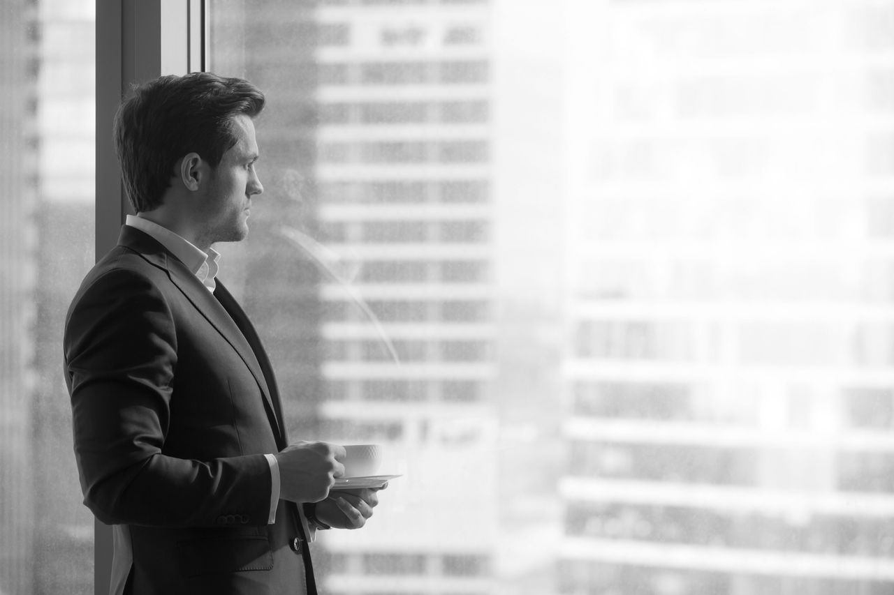 Side view of serious pensive director of prosperous company enjoying cup of coffee in the morning, thoughtful businessman in suit looking through big office window at dawn sunrise city, copy space